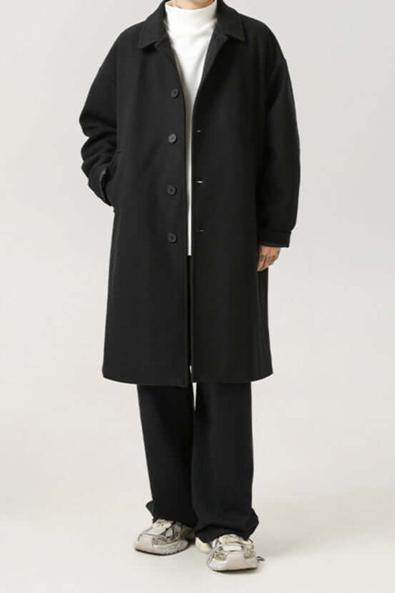 Black oversized button-up coat worn over trousers and trainers, full length
