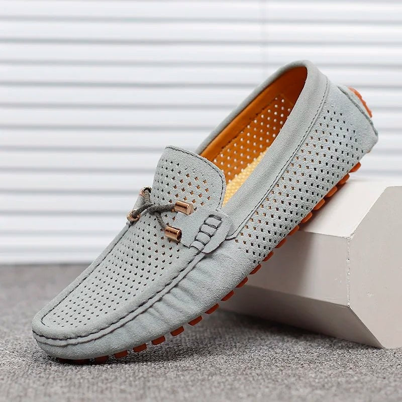 Grey suede perforated loafers with orange sole resting on a white block