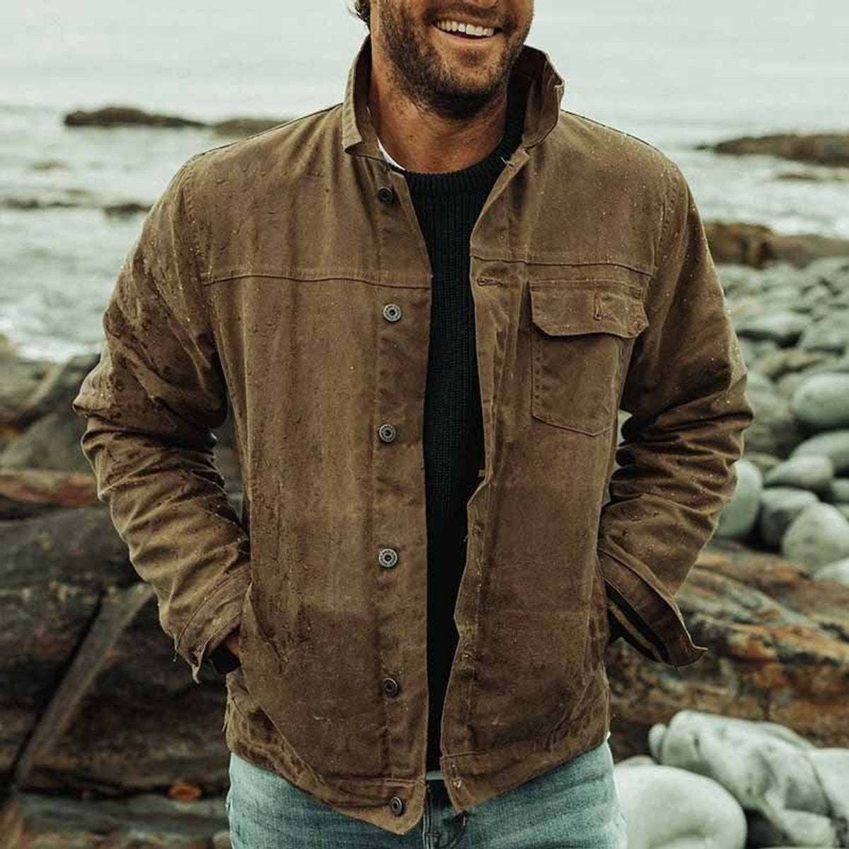 Model smiling in brown button-up outdoor jacket standing on coastal rocks