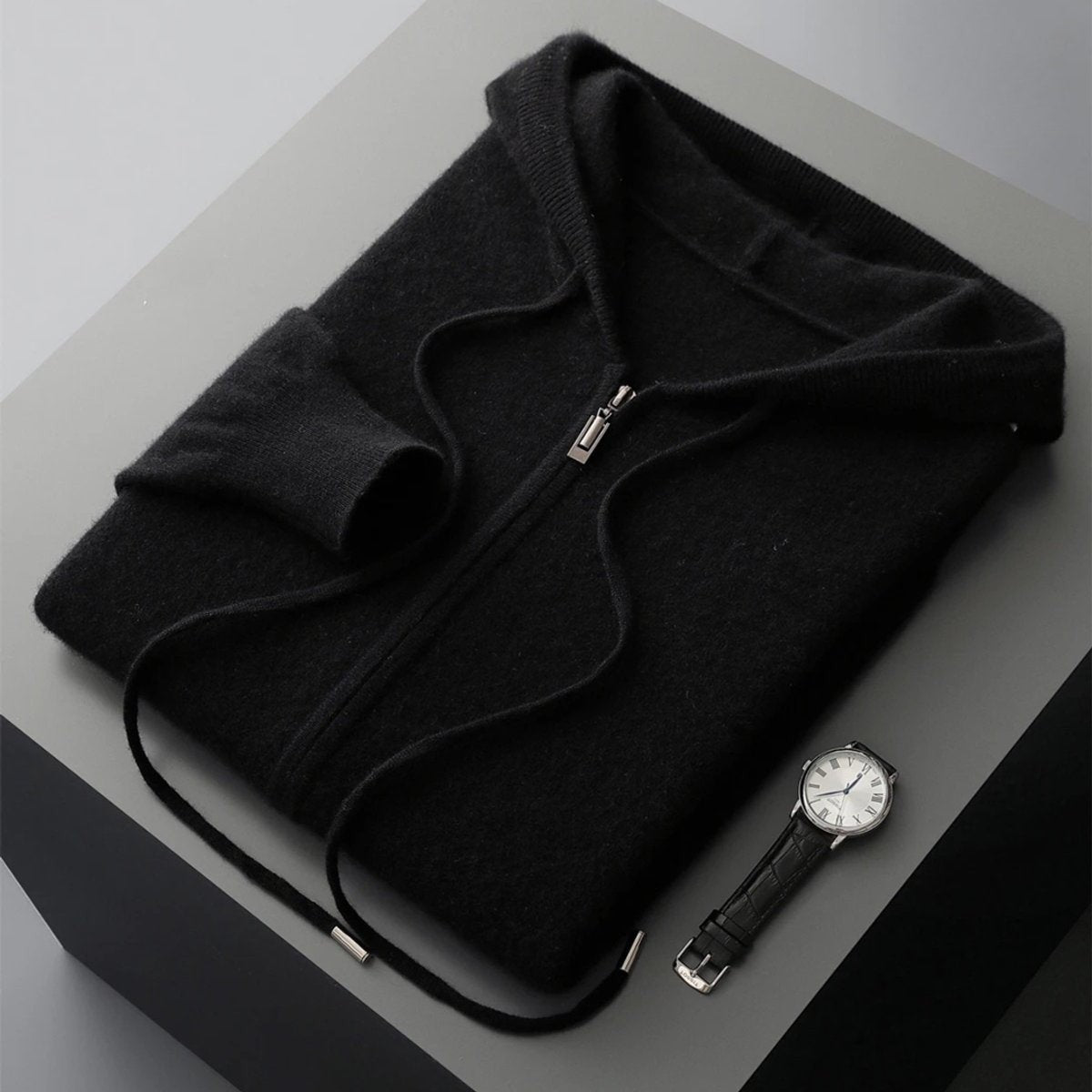 Folded black cashmere knit zip hoodie with ribbed hood beside a leather watch