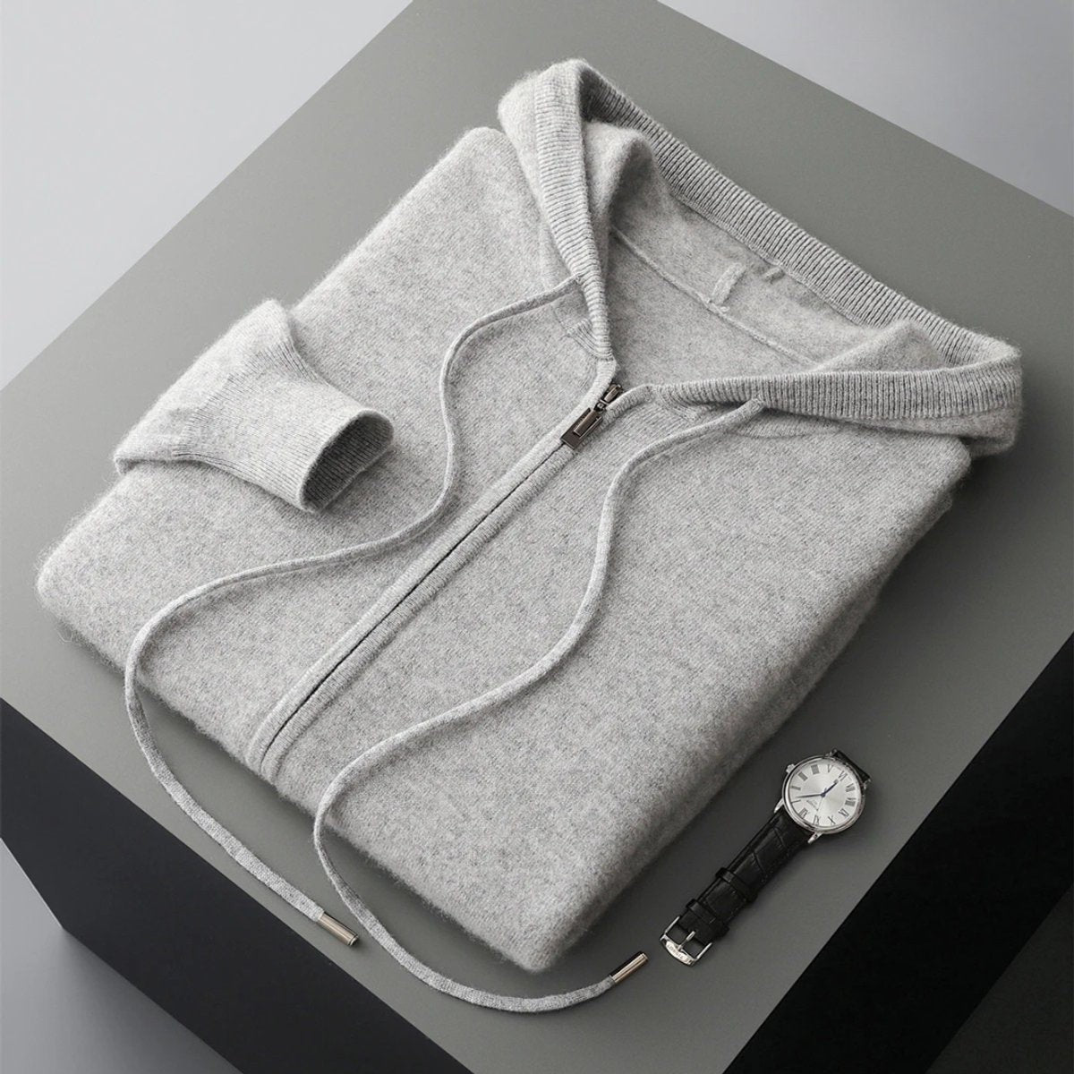 Folded light grey cashmere knit zip hoodie with drawstring hood beside a watch