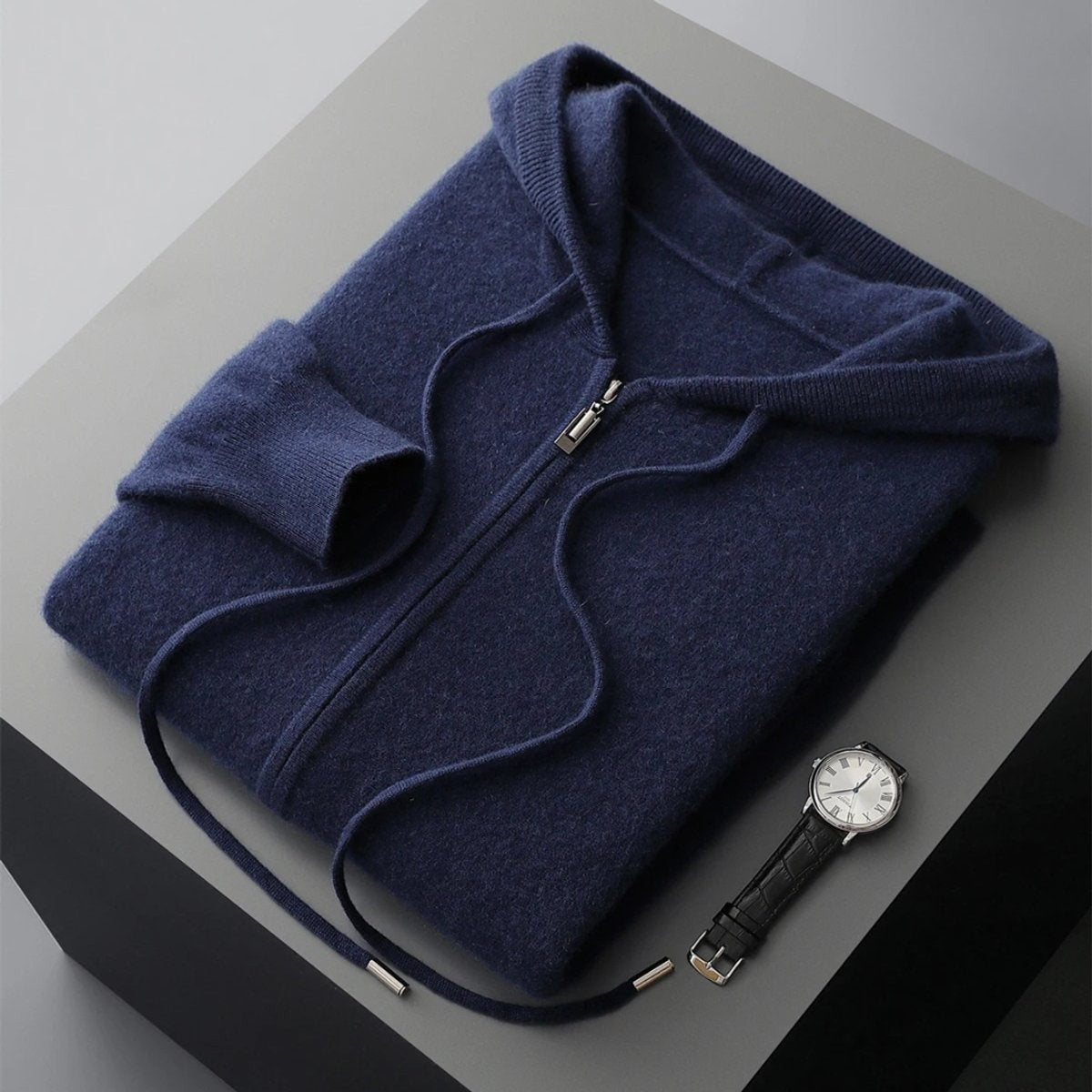 Folded navy cashmere knit zip hoodie with ribbed cuffs beside a leather watch