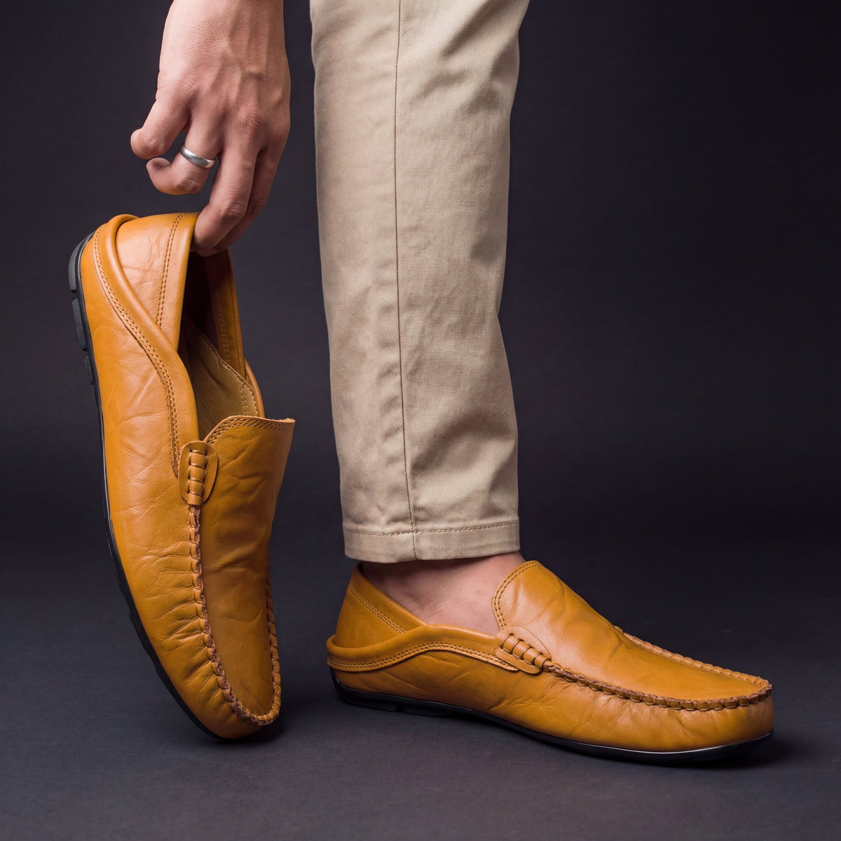 Man holding tan leather moccasin while wearing matching pair on feet