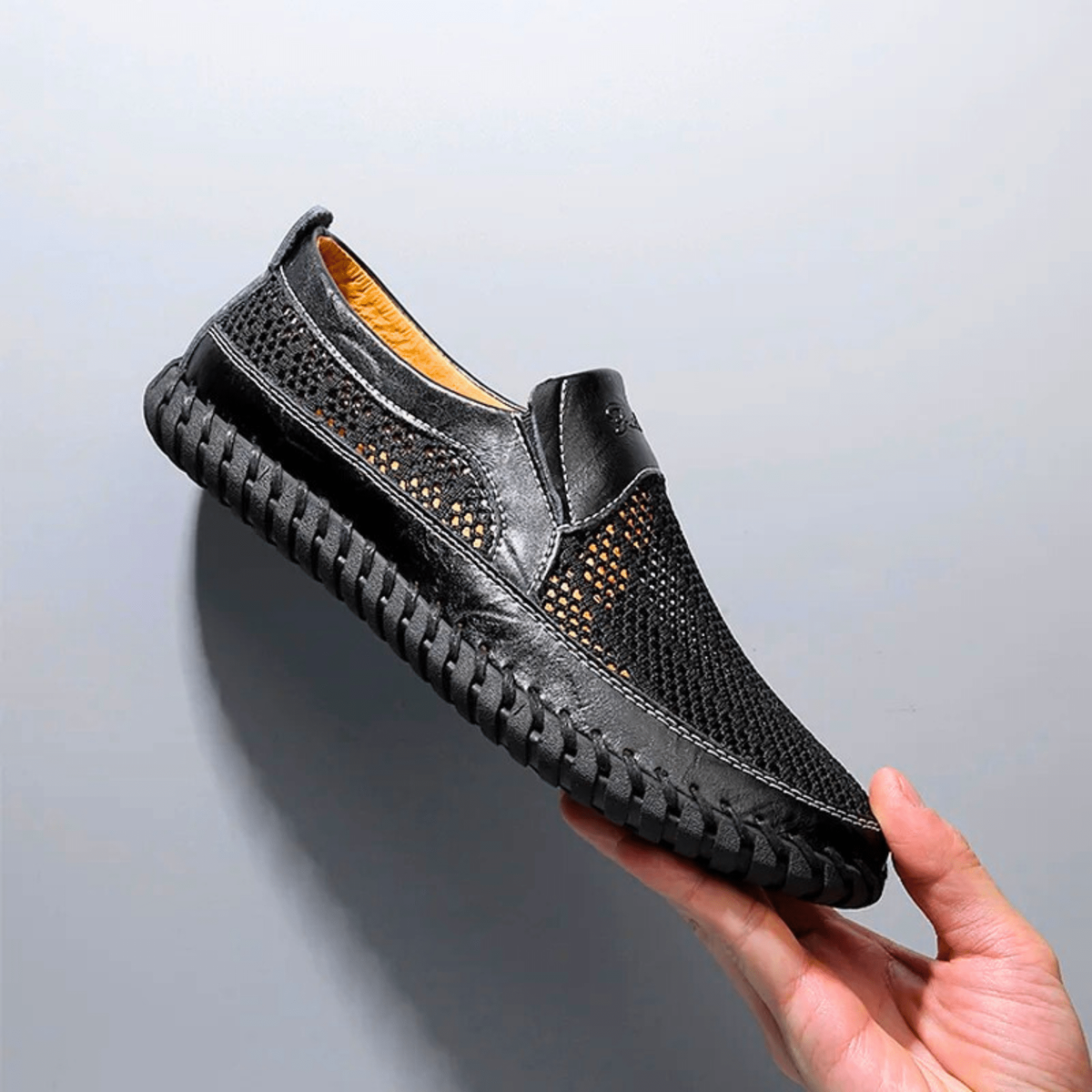Black woven-mesh loafer held up, showing tan leather lining and hand-stitched sole